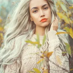 Kobieta z burzą waniliowy blond włosów i pomarańczowymi ustami w beżowym swetrze, otoczona jesiennymi liśćmi.