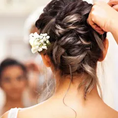 Elegancka fryzura na ślub cywilny z wplecionymi białymi kwiatami. Dłonie stylisty dopinają ostatnie pasma.