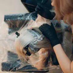 Fryzjerka w czarnych rękawiczkach nakłada farbę na blond włosy. Kluczowe jest, ile trzymać farbę na włosach, by uzyskać pożądany efekt.