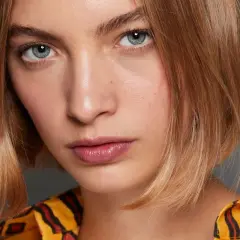 Młoda kobieta z rudymi włosami w stylu long bob, z błękitnymi oczami i błyszczącymi ustami, ubrana w żółtą bluzkę z czarno-czerwonym wzorem.