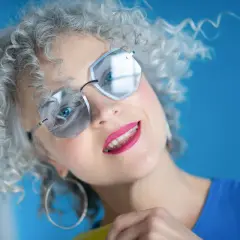 Kobieta z kręconymi włosami po farbowaniu na siwo, w okularach i z wyrazistą szminką.
