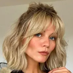 Kobieta z niebieskimi oczami i piegami, z blond włosami upiętymi w luźny kok i stylową grzywką na boki.