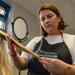 Fryzjerka w fartuchu L'Oréal Professionnel wykonuje podcinanie końcówek blond włosów.