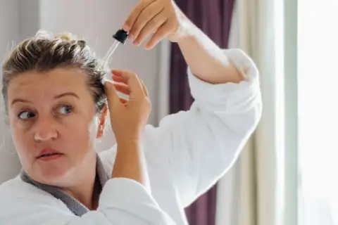 Kobieta w szlafroku aplikuje serum na skórę głowy. Wierzy, że goździki i rozmaryn na włosy przyniosą jej ulgę i wzmocnią kosmyki.