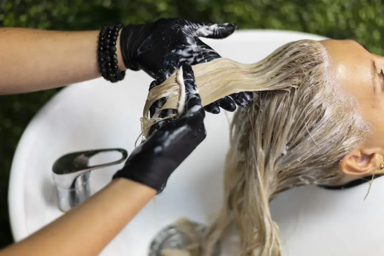 Fryzjerka w czarnych rękawiczkach nakłada produkt na blond włosy, widoczne są siwe odrosty.