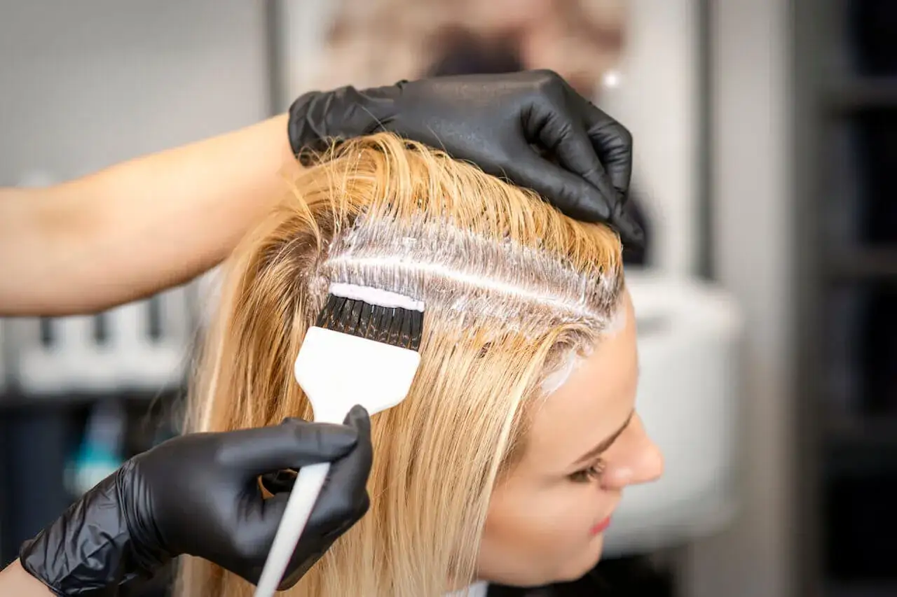 Fryzjerka w czarnych rękawiczkach nakłada rozjaśniacz na blond włosy. Czy myć włosy przed rozjaśnianiem? To pytanie zadaje sobie wiele osób.