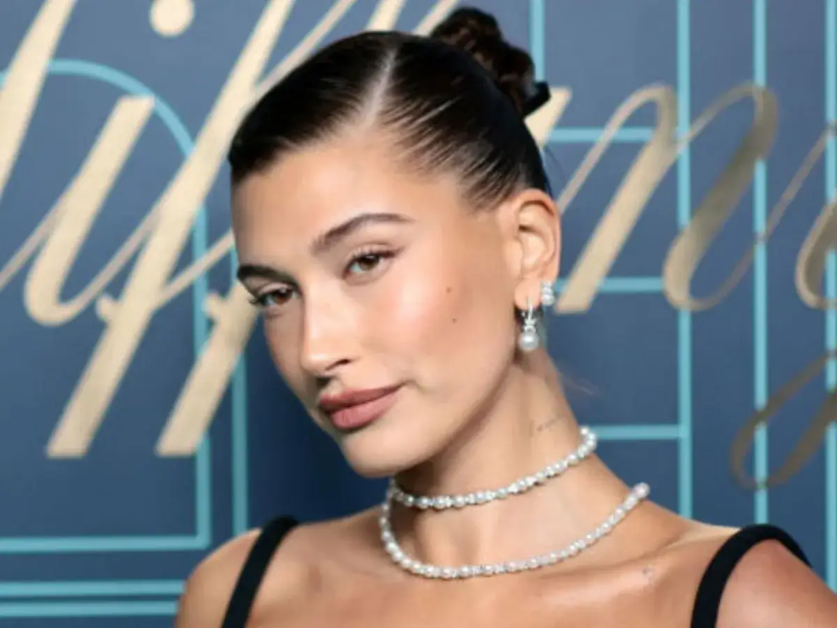 Elegancka fryzura typu sleek bun, perłowa biżuteria i subtelny makijaż podkreślają urodę kobiety na tle błękitnej ściany z logo.