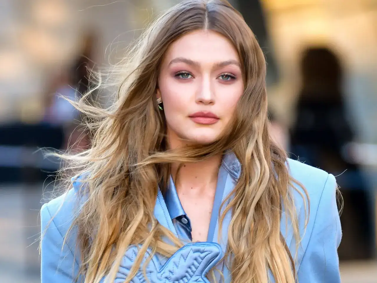Gigi Hadid w błękitnej marynarce, jej blond włosy falują, a słońce rozjaśnia je, tworząc złote refleksy.