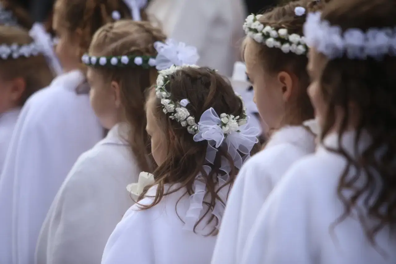 Dziewczynki w białych strojach, z delikatnymi fryzurami komunijnymi ozdobionymi wiankami z białych kwiatów i wstążkami.